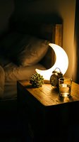 Cozy bedroom nightstand with glowing crescent moon lamp, succulent plant, vintage alarm clock and candle creating warm ambient lighting atmosphere.