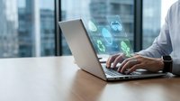 Businessman working on laptop with digital security icons floating above keyboard in modern office with city skyline view. Cybersecurity and data protection concept.