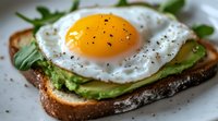Sunny side up egg on avocado toast garnished with fresh arugula, black pepper and seasonings on rustic bread, close up view of breakfast or brunch meal.