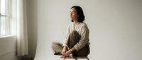 Young Asian woman in casual beige outfit sitting cross-legged on floor in minimalist room with natural window light for meditation and wellness content.