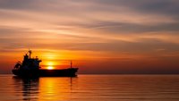 Cargo ship silhouette sailing on calm ocean waters during golden sunset with dramatic orange and pink sky clouds reflecting on water surface.