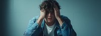 Young Caucasian man in denim jacket showing signs of depression and anxiety, holding head in hands against turquoise wall, dramatic panoramic shot.