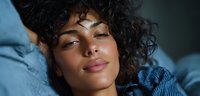 Young woman with curly hair relaxing on blue bedding, eyes half-closed in peaceful contemplation, soft lighting creating intimate atmosphere.