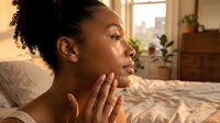 Young African American woman applying skincare routine in bright bedroom with natural morning light streaming through window creating warm atmosphere.