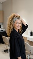 Smiling middle-aged Caucasian woman with curly blonde and gray hair posing in a modern hair salon wearing a black robe.