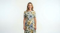 Middle-aged Caucasian woman wearing floral print dress with blue and yellow flowers on cream background, short sleeves, standing against white backdrop for fashion and lifestyle use.