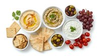 Overhead view of Mediterranean appetizer spread with hummus, tzatziki, olives, grapes, tomatoes, pita bread and crackers on white background. Perfect for food styling, restaurant menus, catering and culinary content.