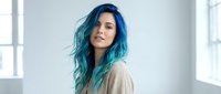 Young woman with vibrant blue turquoise hair in casual beige sweater standing by window in bright modern interior space for lifestyle and fashion concepts.