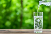 Pouring fresh pure water from bottle into drinking glass on wooden table outdoors with green natural bokeh background. Refreshing summer beverage concept.