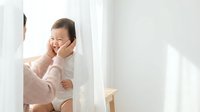 Happy Asian toddler girl in cozy knit sweater playing peek-a-boo behind white curtains in bright sunlit room for family lifestyle content.