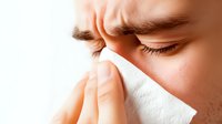 Close-up of person holding tissue to nose, showing signs of nasal congestion, cold or allergy symptoms against light background, medical health concept.