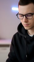 Young Caucasian male wearing black hoodie and eyeglasses, looking downward with contemplative expression under neon light, suitable for youth, technology, and lifestyle concepts.