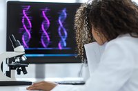 African American female scientist in lab coat analyzing DNA structure on computer screen while working with microscope in modern research laboratory setting.