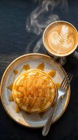 Fluffy pancakes drizzled with honey spelling sweet, served with steaming latte art on dark slate for a cozy breakfast concept.