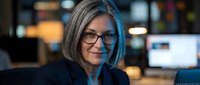 Middle-aged woman with gray hair and eyeglasses wearing dark blazer in modern office environment with computers and blurred background lighting.
