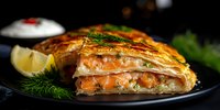 Flaky puff pastry filled with fresh salmon, cream cheese, and herbs, sliced to show layered filling, served on black plate with lemon and dill garnish.