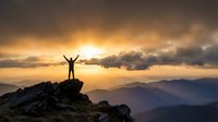 Silhouette of person with raised arms celebrating success on mountain peak at golden sunrise with dramatic cloudy sky and layered mountain ranges.