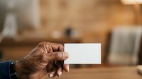 Mature man with dark skin tone holding blank white business card in hand against wooden workshop background with tools and equipment.