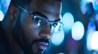 Young African American man in glasses illuminated by blue neon lights at night, close-up portrait with bokeh background showing contemplative expression.