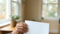 Senior woman with wrinkled hands holding blank white paper at home near window with natural light, suitable for communication, messaging, announcements.