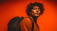 Young African American man with curly hair wearing orange sweater against vibrant red background. Modern portrait photography for fashion and lifestyle projects.