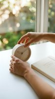 Person holding wooden alarm clock at white desk with open notebook near window with garden view, morning routine and time management concept.
