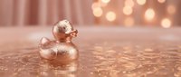 Elegant rose gold rubber duck toy with glittery finish sits on reflective surface with warm bokeh lights creating luxurious bathroom decor atmosphere.