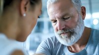 Senior Caucasian man with white beard and gray t-shirt listening intently to female medical professional during healthcare consultation in modern clinic setting.