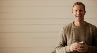 Young Caucasian man with blonde hair, wearing a beige cable-knit sweater, smiling while holding a smartphone against neutral background for lifestyle and technology concepts.