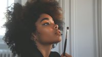 Young African American woman with natural afro hair and blue eyeshadow in profile view during makeup application with brush against soft white background.