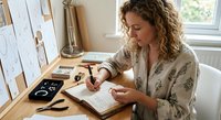 Young Caucasian woman with curly hair writing in notebook at wooden desk in bright home office workspace with calendar and supplies.