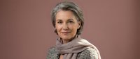 Elegant mature Caucasian woman with gray hair wearing beige scarf and knitted sweater smiling warmly against soft pink background for lifestyle content.