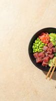 Poke bowl with raw tuna cubes, edamame, cucumber, ginger, sesame seeds and wooden chopsticks on beige surface. Asian cuisine meal preparation.