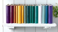 Colorful hardcover books arranged in rainbow spectrum order standing in row on white wooden shelf against light wall with green plant decoration.