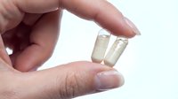 Hand holding two white capsules between fingers on clean background for healthcare and pharmaceutical design applications.