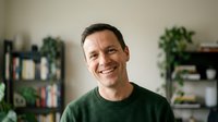 Smiling middle-aged Caucasian man in green sweater at home with plants and shelves in background for lifestyle and wellness content.