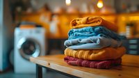 Stack of neatly folded colorful laundry on wooden table with washing machine in background, warm ambient lighting creates cozy laundry room atmosphere.