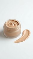 Open glass jar of beige foundation cream with smooth swirl texture and makeup swatch on white background for cosmetic product photography and beauty advertising.