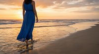 Woman in blue maxi dress walking barefoot on sandy beach at golden sunset with ocean waves, peaceful evening stroll by the sea for relaxation and wellness.