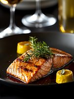 Grilled salmon fillet garnished with fresh rosemary and lemon on black plate. Golden-brown glazed fish with crispy skin and herbs creates elegant restaurant atmosphere.