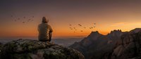 Lone person in a hoodie sitting on a rocky mountain cliff at sunset, watching birds fly over a dramatic orange sky and misty peaks.