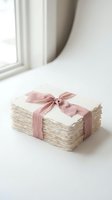 Stack of cream-colored papers tied with dusty rose ribbon bow, displayed on white surface near window with natural light for gift wrapping and stationery concepts.