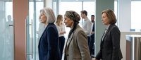 Mature women professionals in business casual attire conversing in a modern office corridor with colleagues in background, suitable for corporate, diversity, and workplace communication concepts.