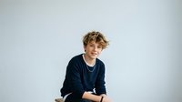 Young male with blonde wavy hair and fair complexion wearing dark navy sweater and silver chain necklace, smiling at camera against white background for portrait photography.