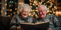 Elderly couple sharing memories while reading photo album together during holiday season with Christmas lights in background.