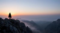 Silhouetted hiker standing on rocky mountain peak at sunrise with misty valleys below, conveying adventure, exploration and outdoor achievement for travel and lifestyle content.