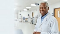 Senior African American male doctor in white coat smiling confidently in modern hospital corridor with medical equipment and bright lighting.