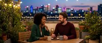 Young African American woman and Caucasian man enjoying evening conversation on urban rooftop terrace with city skyline backdrop and string lights.