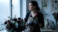Young Caucasian female florist arranging dark burgundy flowers in bright workshop studio. Professional woman creating floral bouquet by window.