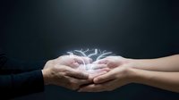 Hands of two people cupping glowing light energy between palms against dark background, representing connection, transfer of power, healing, teamwork, and spiritual exchange.